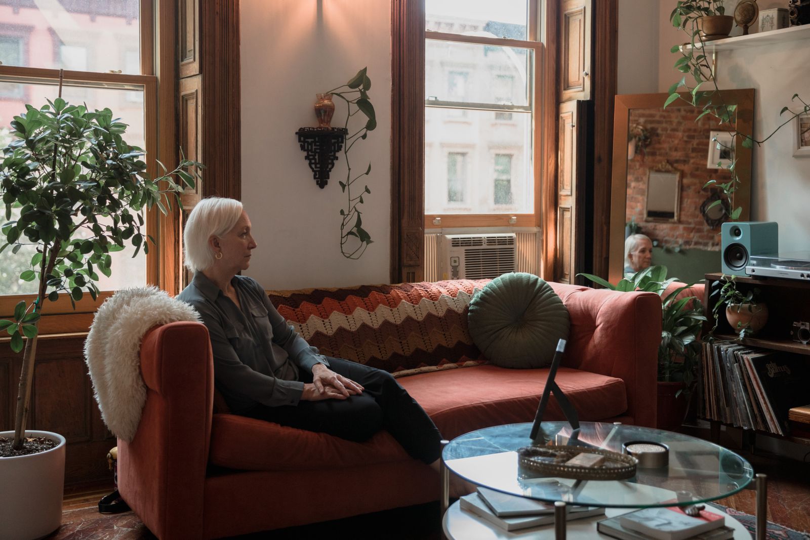 An older woman sitting alone on a red sofa in a sunlit living room, looking toward the window
