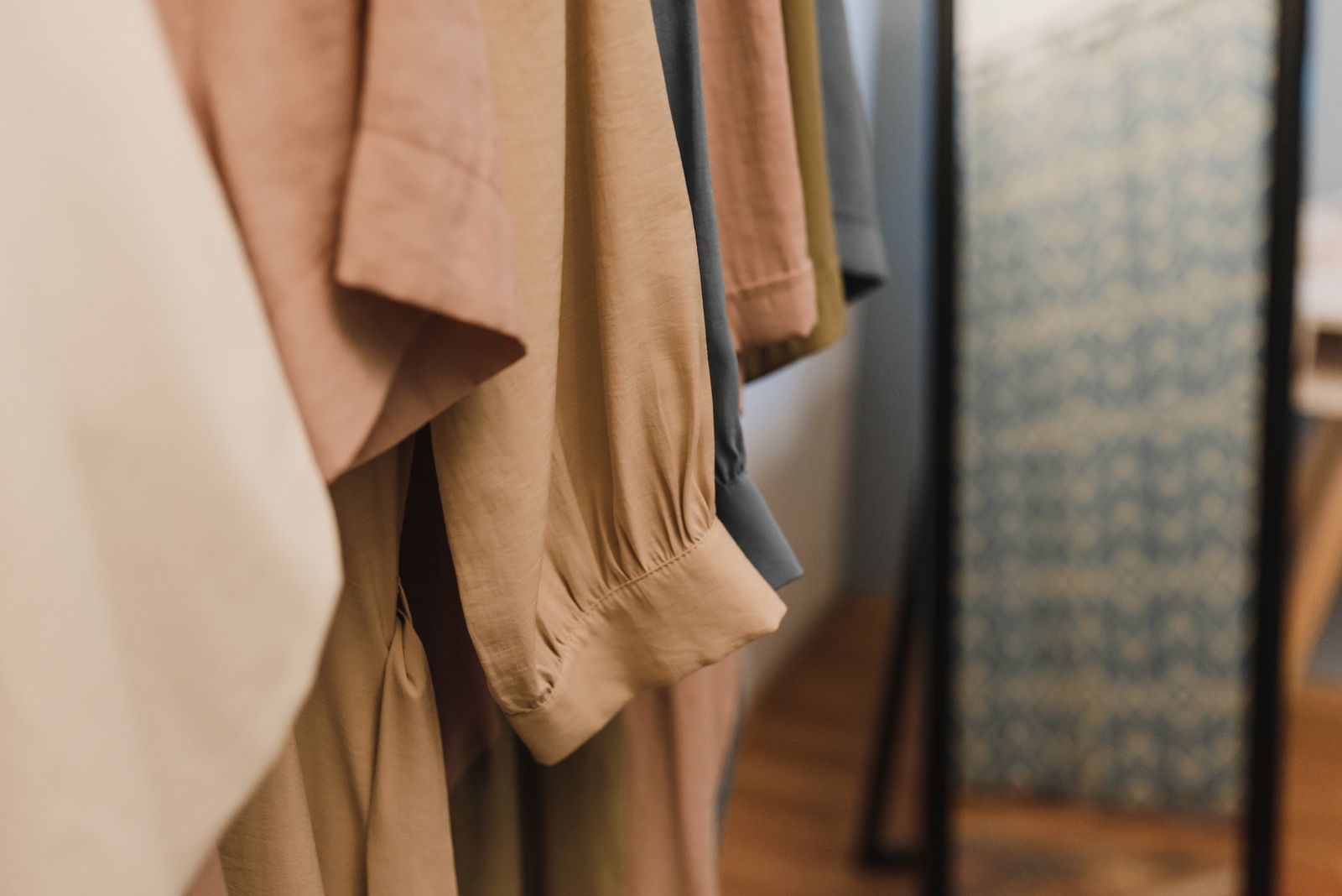 A close view of soft linen clothes in cream, dusty pink, tan and grey hanging on an open rail beside a tall mirror in a quiet, sunlit room