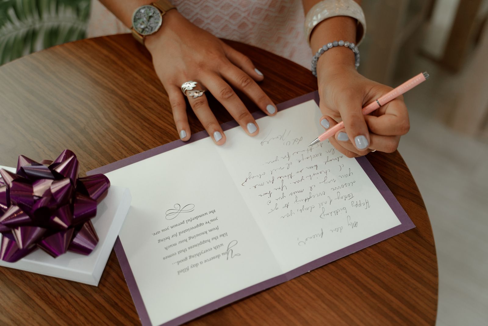 A woman writing a message inside a greeting card on a round wooden table beside a gift bow