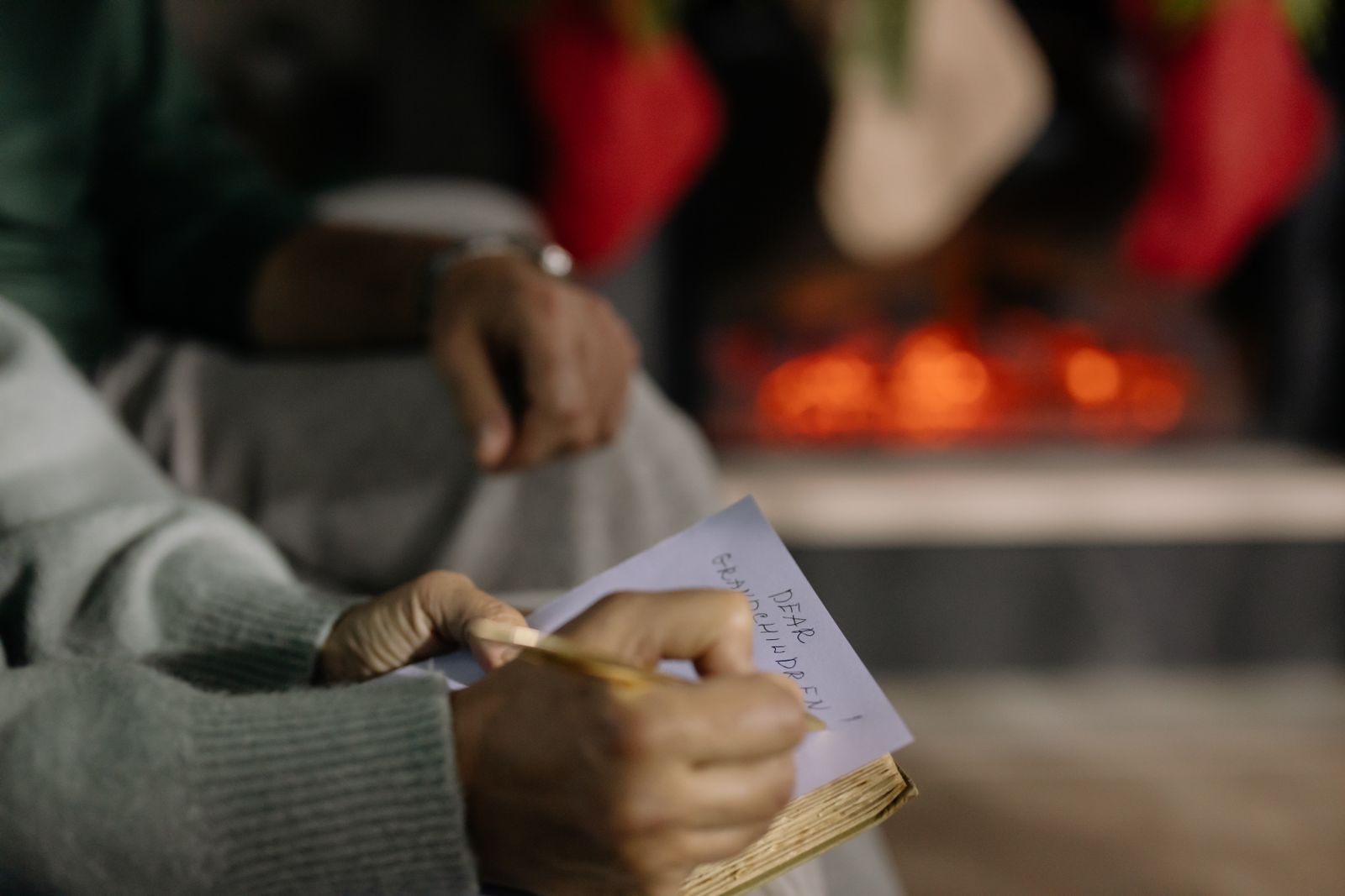 A person in a grey sweater writing a note beside a glowing fireplace hung with stockings