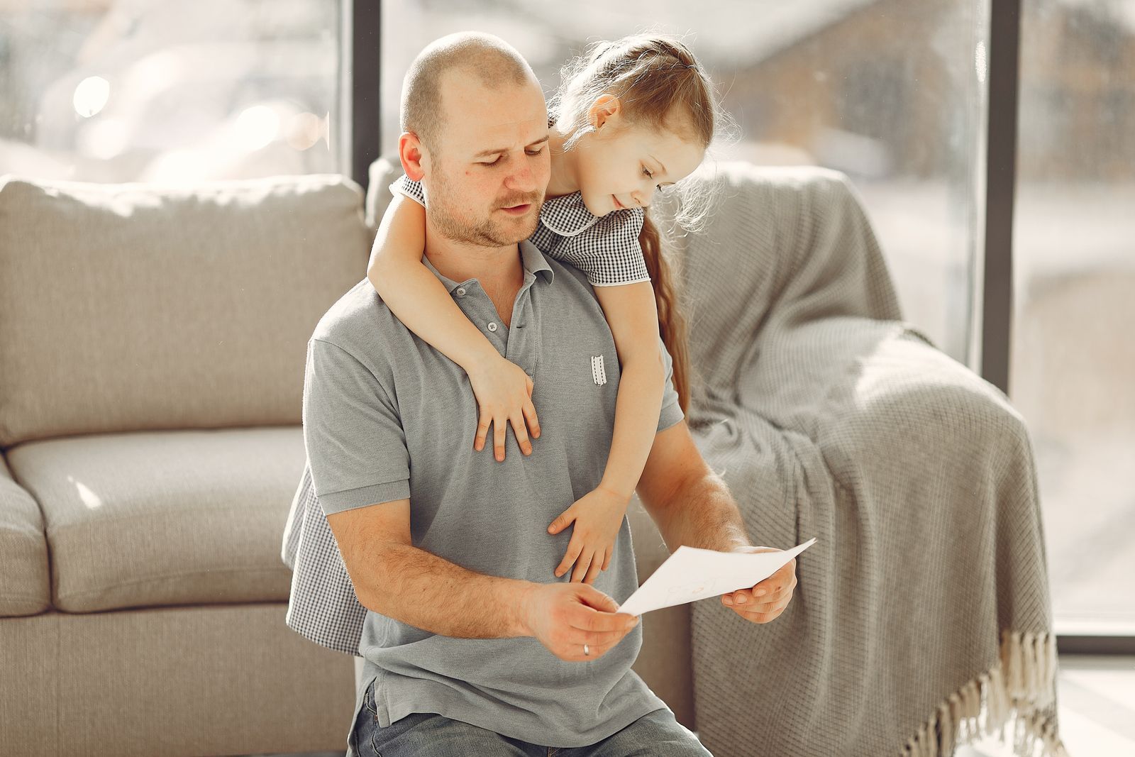 A father on a sofa reading a letter while his young daughter hugs him from behind