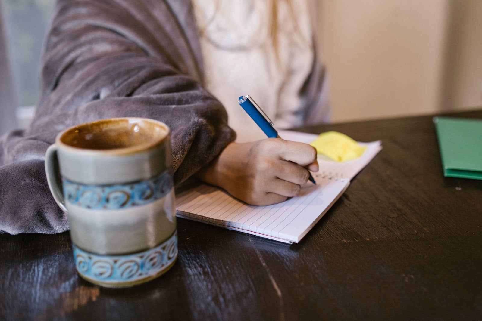 شخص يرتدي رداءً مريحًا يكتب قائمة في دفتر ملاحظات إلى جانب كوب قهوة من السيراميك