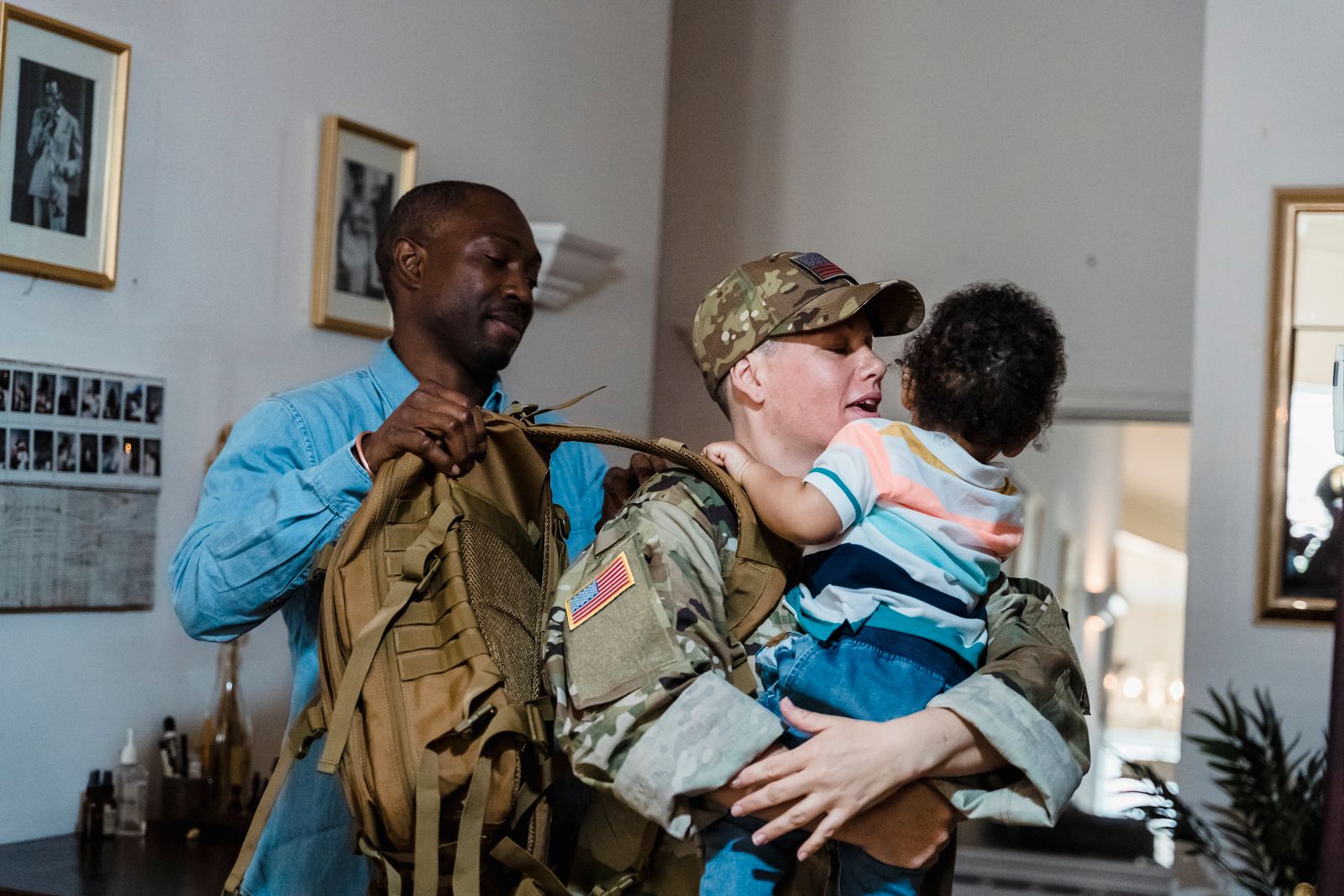A soldier in uniform holding a child while a partner helps lift a packed rucksack