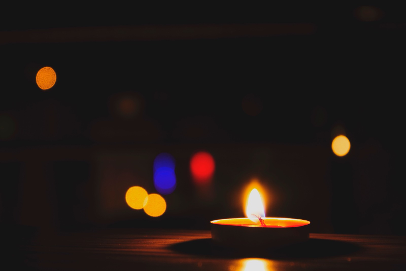 Ein einzelnes brennendes Teelicht leuchtet warm auf einem Holztisch vor weichen, bunten Bokeh-Lichtern