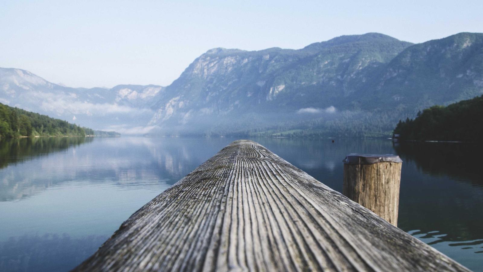 Ein verwitterter Holzsteg führt über einen stillen See auf Nebel und ferne Berge zu