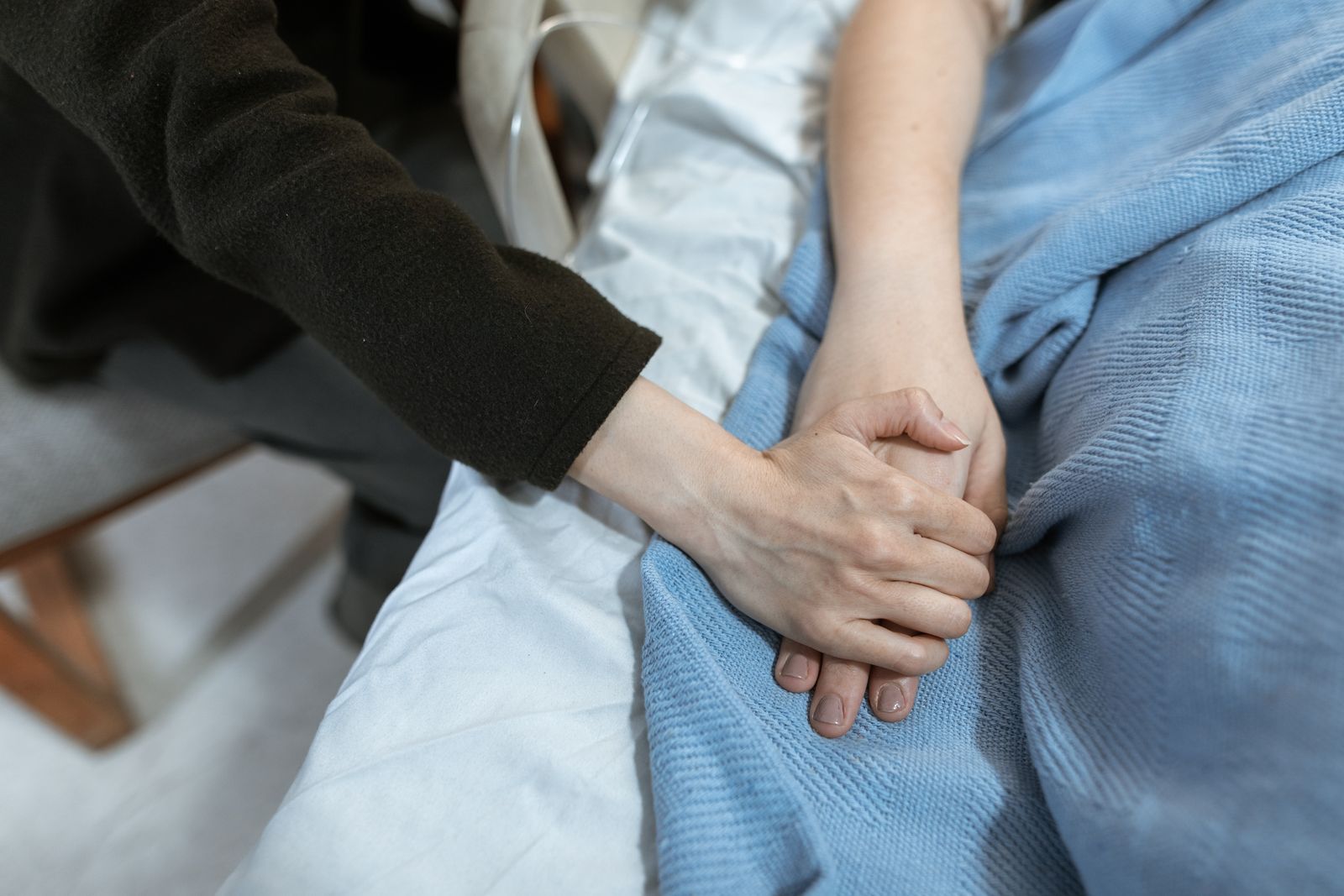 Two hands gently holding another hand resting on a pale blue hospital blanket