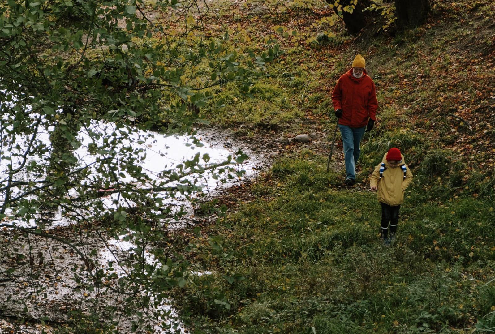 A grandfather in a red jacket and a child in yellow walking together beside an autumn stream