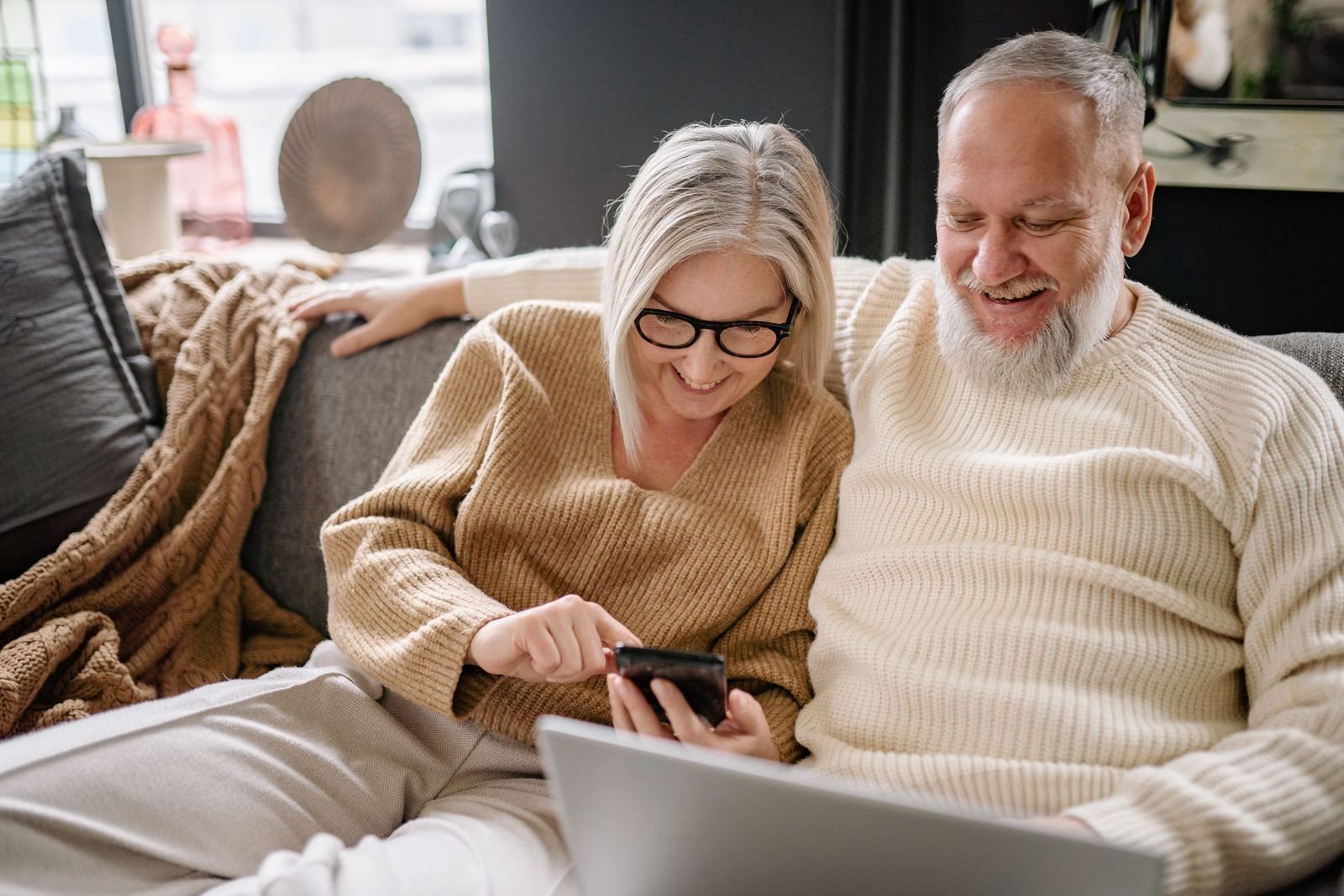 An older couple in knitted jumpers sitting close together on a sofa, both smiling at something on her phone