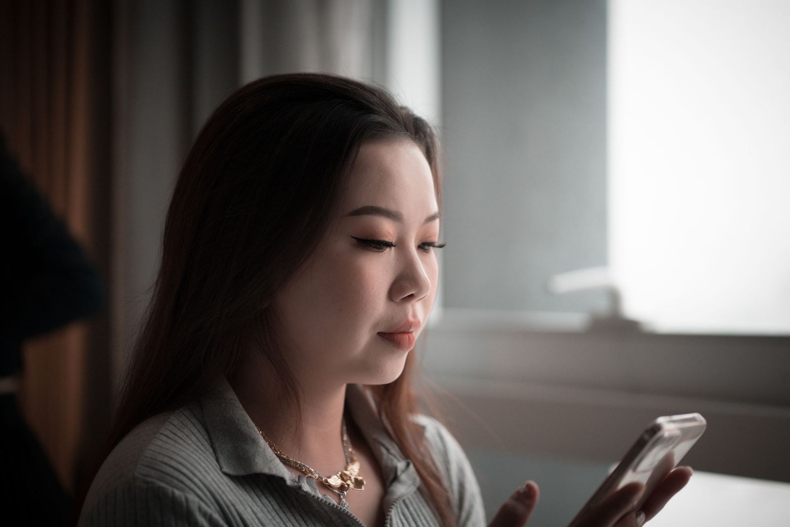 窓からのやわらかな光の中で、うつむいてスマホを見ている若い女性