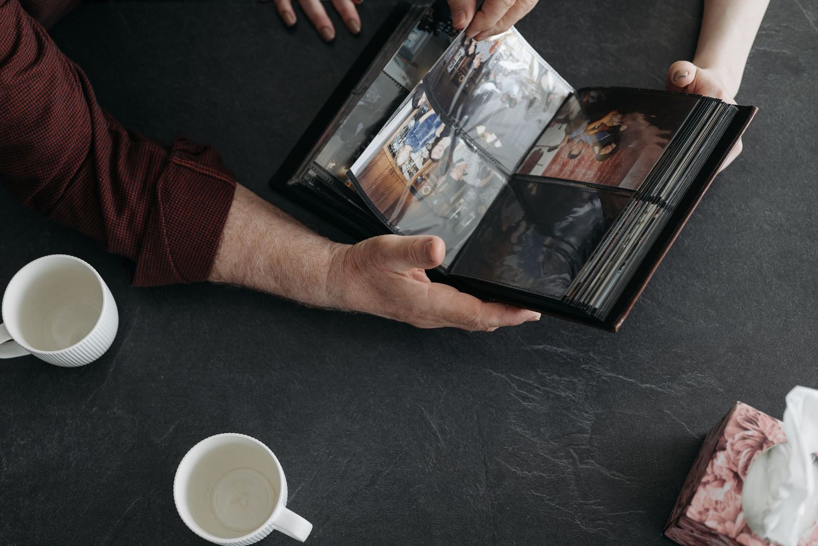 Hands holding open a family album of snapshots over a dark table, with two empty mugs and a box of tissues nearby