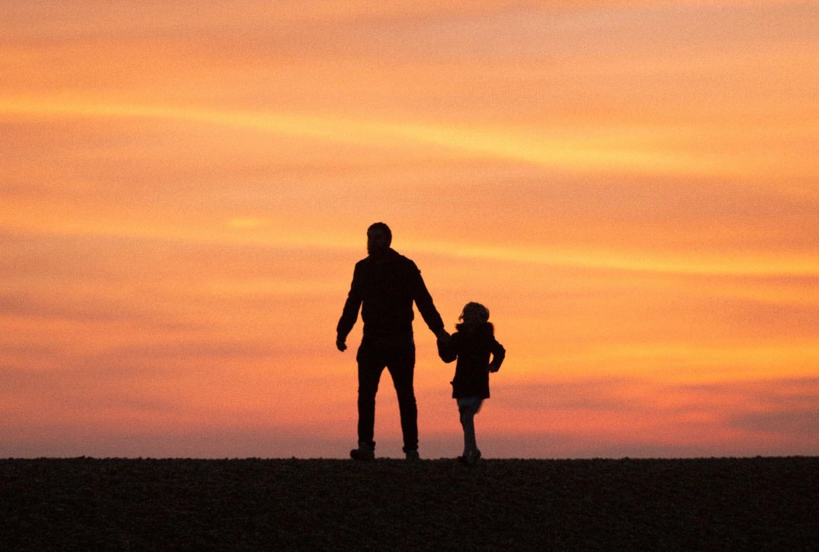 Silhouette eines Elternteils mit kleinem Kind, Hand in Hand vor einem orangefarbenen Abendhimmel