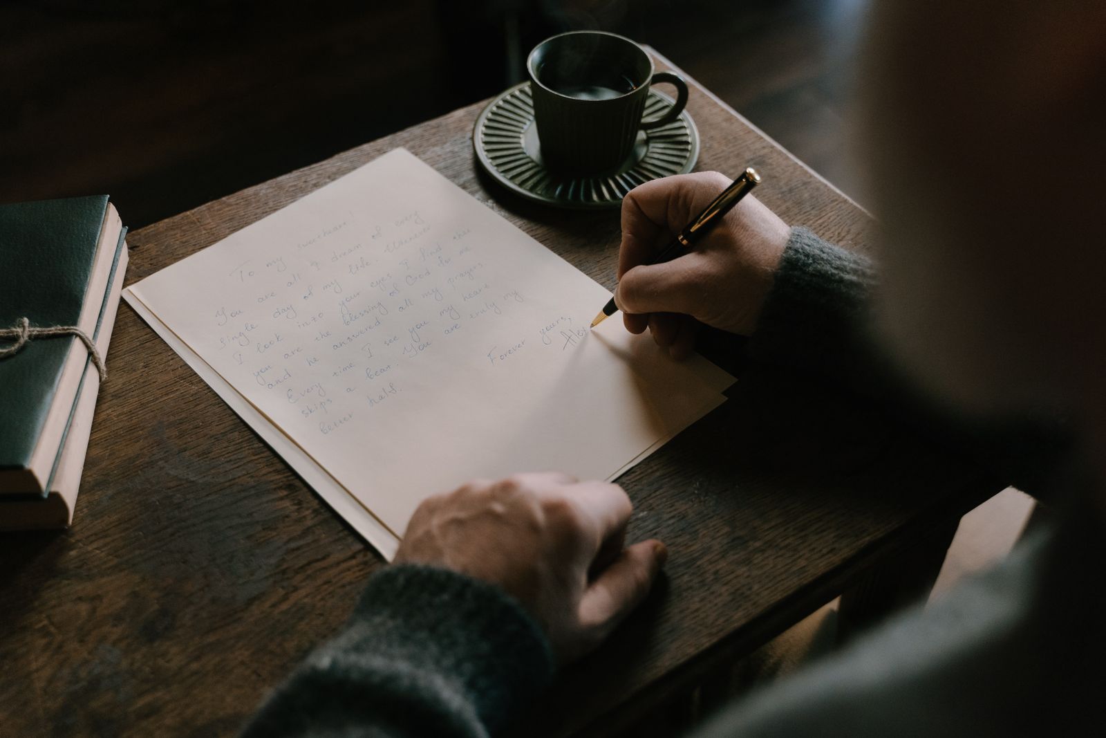 A person writing a letter by hand at a dark wooden desk beside a cup of coffee and a leather notebook
