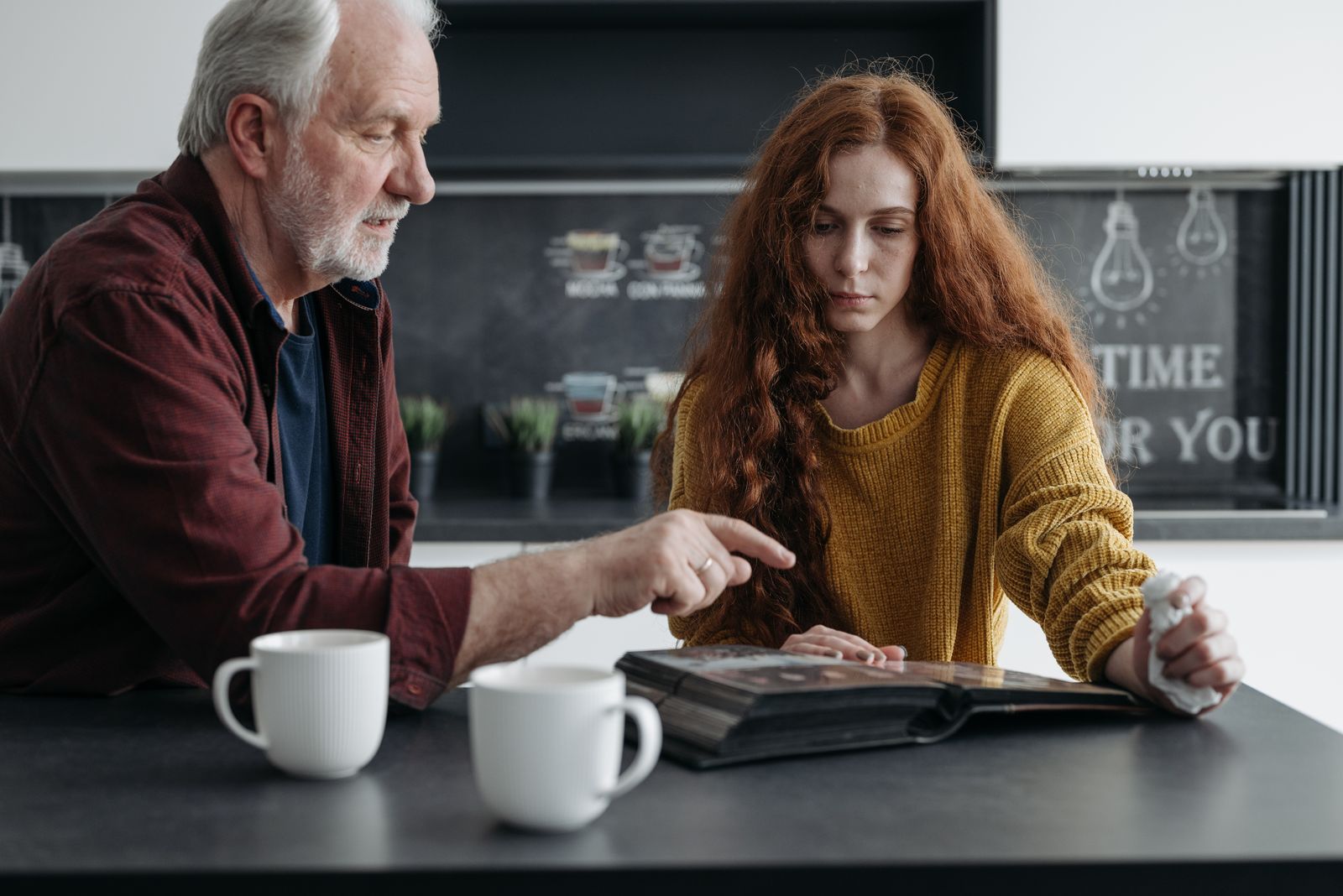 An older man and a young woman at a kitchen table with two mugs, looking through an open family album