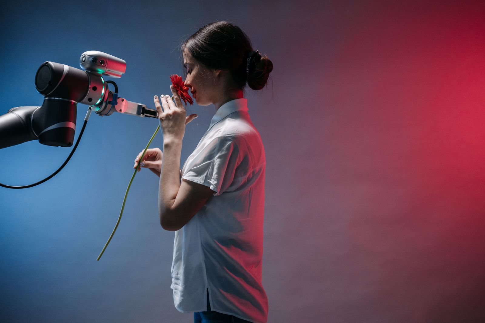 A woman smelling a red flower held out to her by a robotic arm against a blue and red backdrop