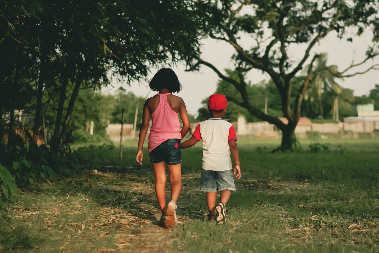 A girl and a younger boy in a red cap walking hand in hand along a grassy path, seen from behind