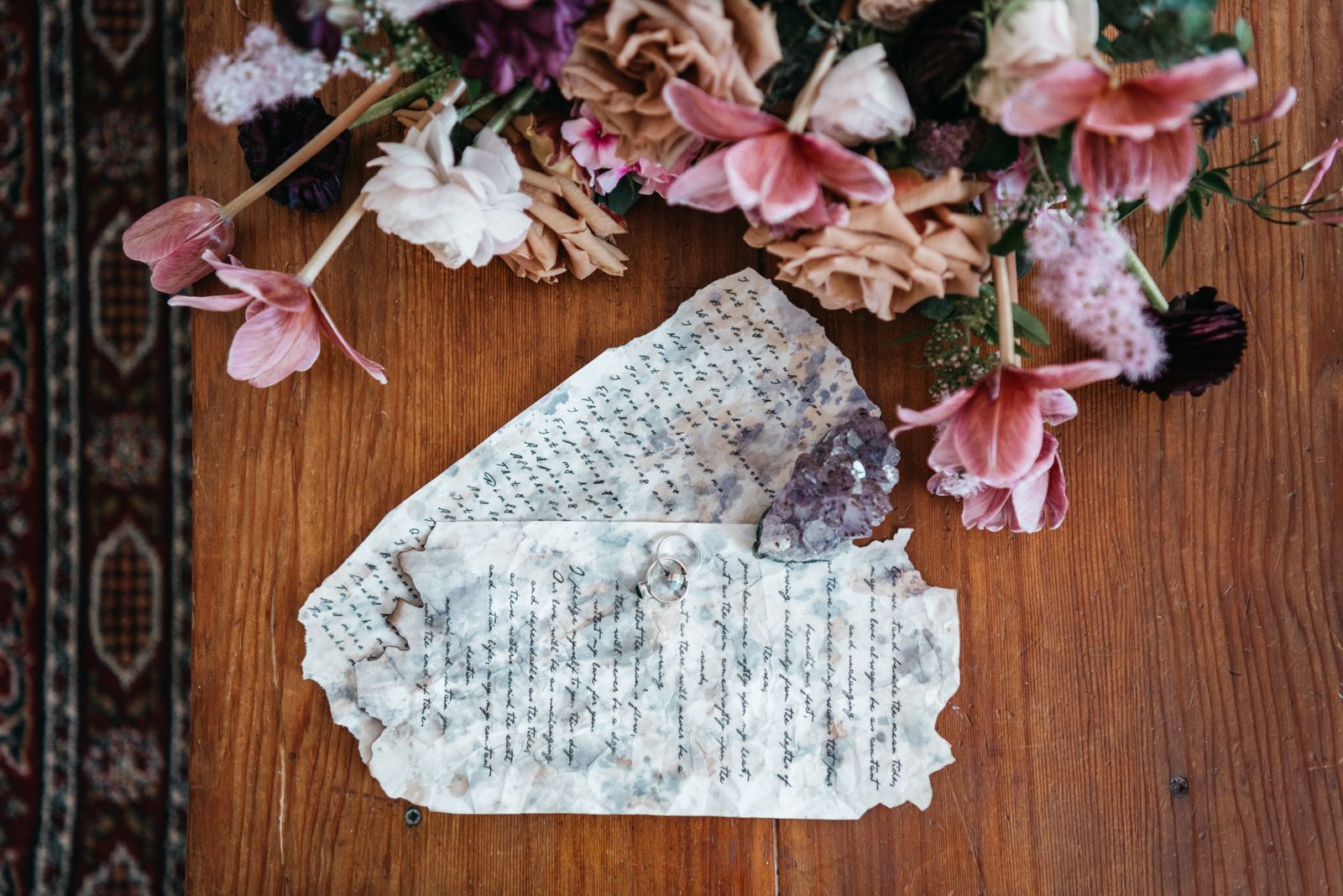 Two wedding rings resting on handwritten letter pages laid out on a wooden table beside flowers